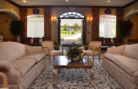 A cozy living room area with two beige sofas and two armchairs arranged around a wooden coffee table with a floral centerpiece. The room has patterned carpet, large windows with curtains, wall sconces, and an open door leading outside to a garden and a building in the background.