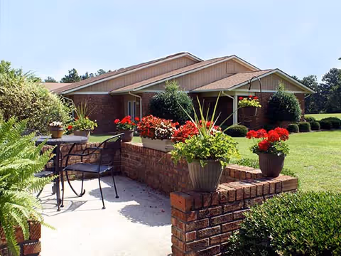 Outdoor patio area with a small round table and two metal chairs surrounded by brick planters filled with various green plants and red flowers. In the background, there is a single-story building with a sloped roof and well-maintained lawn and shrubs.