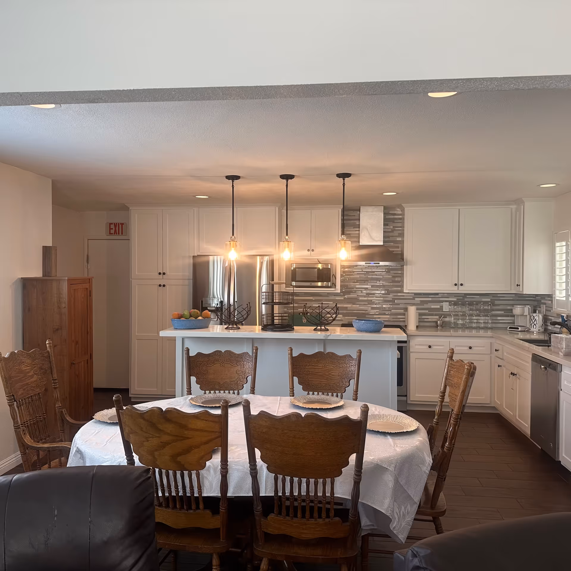 A bright kitchen and dining area with a round table covered with a white tablecloth and six wooden chairs. The kitchen features white cabinets, a stainless steel refrigerator, microwave, and dishwasher, a tiled backsplash, and three hanging pendant lights above a white island counter. There is a wooden cabinet and an exit sign visible in the background.