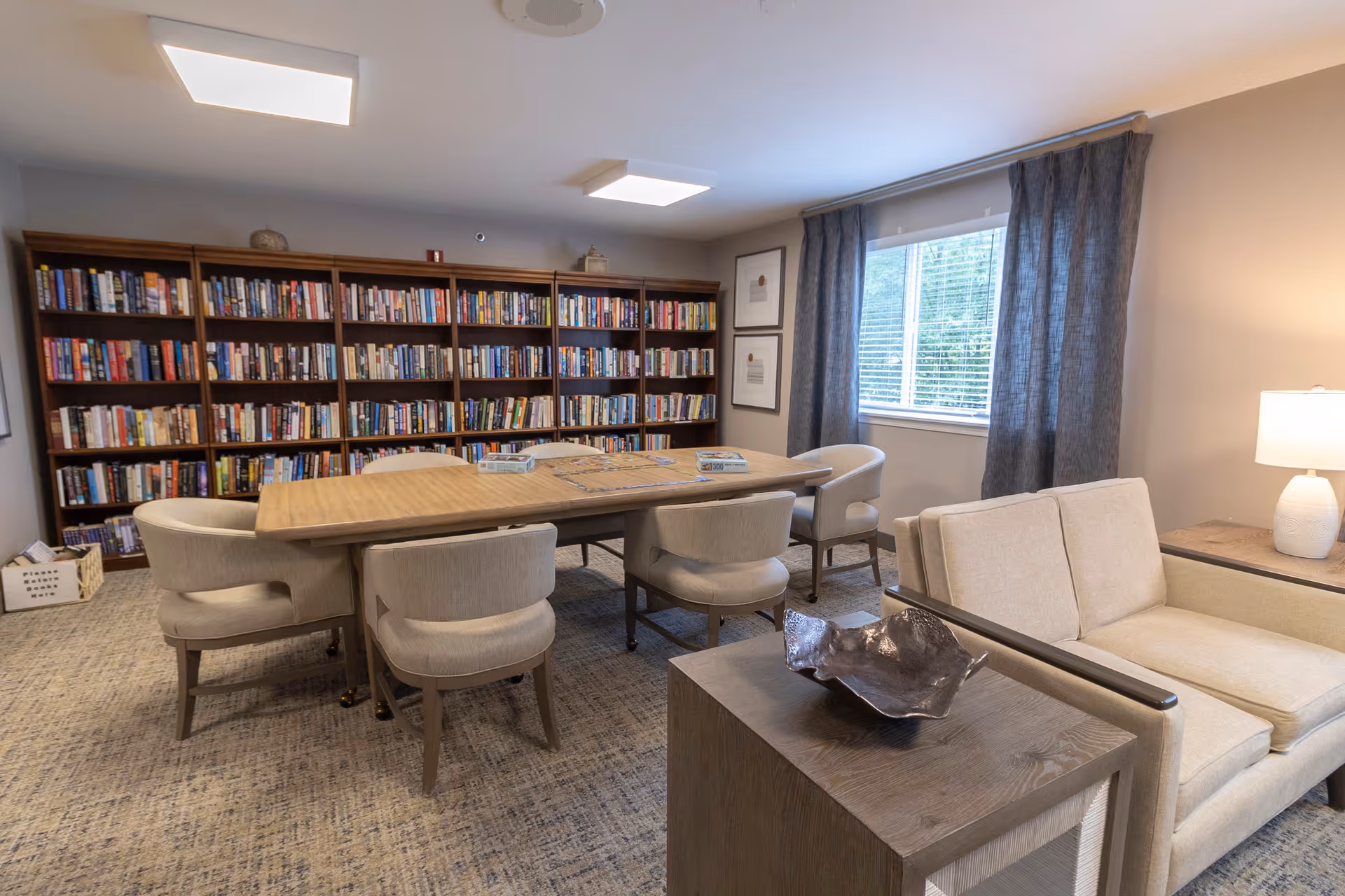 A cozy library room with a large wooden table surrounded by six cushioned chairs. Behind the table is a tall bookshelf filled with books. To the right, there is a window with gray curtains and a beige loveseat next to a wooden side table with a white lamp. The room has carpeted flooring and soft overhead lighting.