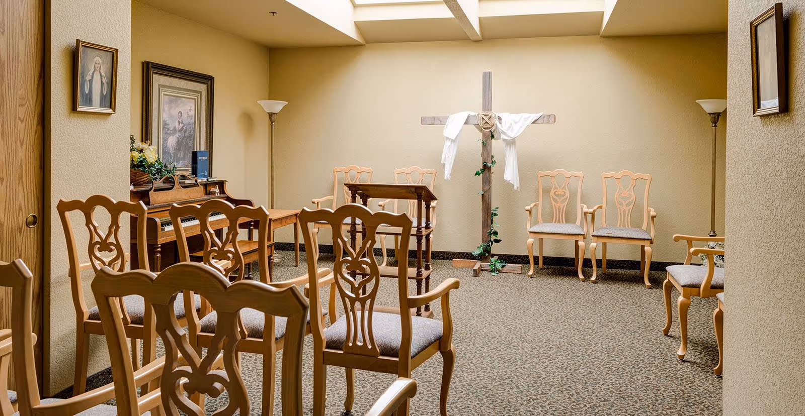 A small chapel room with wooden chairs arranged in rows facing a wooden podium and a large wooden cross draped with white cloth and decorated with greenery. There is a piano against the left wall with framed religious artwork above it, and two floor lamps in the corners of the room. The walls are beige and the carpet is patterned.