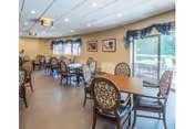 A dining room with multiple wooden tables and cushioned chairs arranged neatly. The room has large windows with blue valances allowing natural light to enter, and framed artwork on the beige walls. The ceiling has recessed lighting and a decorative light fixture.