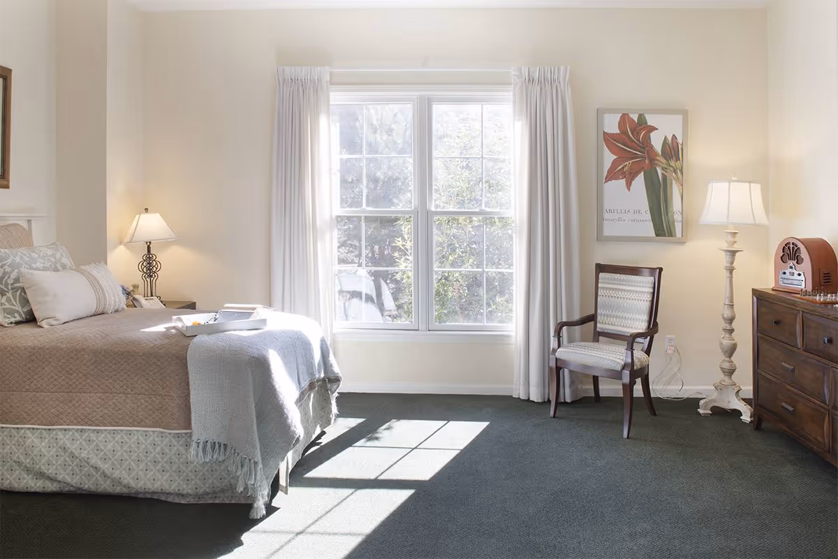 Bright carpeted bedroom with a bed, bedside lamps, a window with curtains, an armchair, and a wooden dresser.