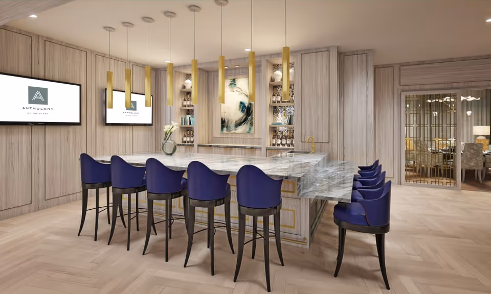 Modern interior space featuring a large marble island with eight blue cushioned bar stools. The walls are covered with light wood paneling, and two flat-screen TVs display the logo 'Anthology of the Plaza.' Above the island, there are multiple gold pendant lights. Shelves with decorative items and a piece of abstract art are visible in the background. To the right, there is a glass door leading to another room with dining chairs and tables.