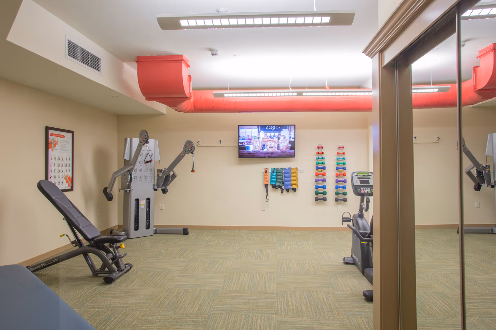 A fitness room with exercise equipment including a weight bench, a multi-function machine, a stationary bike, and racks of colorful dumbbells and ankle weights. A flat-screen TV is mounted on the wall, and a large mirror reflects part of the room. The ceiling has red ventilation ducts and fluorescent lighting.
