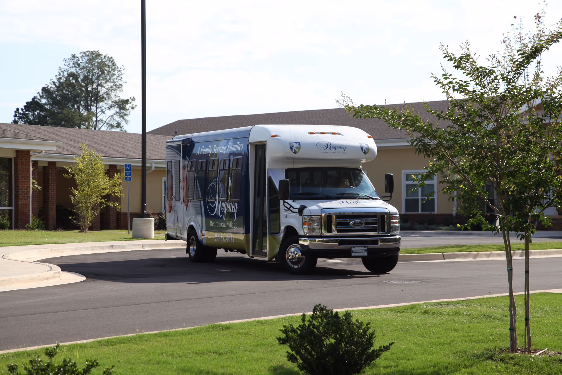 A white and blue shuttle bus with the Regency Retirement Village logo parked on a driveway in front of a single-story building with a brown roof. There are small trees and green grass around the driveway.