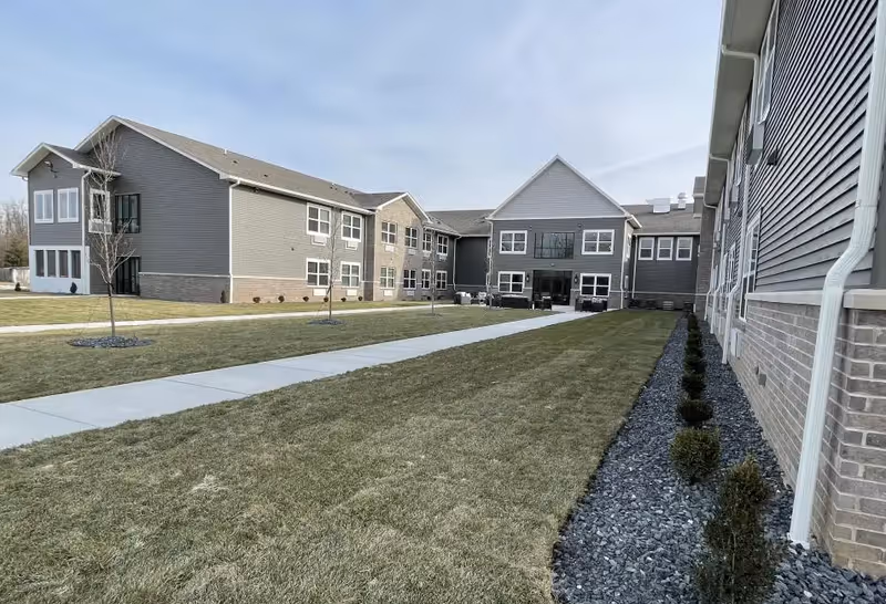 Exterior view of a modern assisted living facility with gray siding and brick accents. The building surrounds a grassy courtyard with a concrete walkway and small trees planted along the path. The sky is clear and the area is well-maintained.