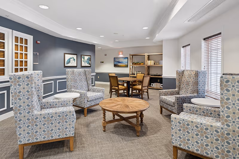 A cozy common area in an assisted living facility featuring four patterned armchairs arranged around a round wooden coffee table. In the background, there is a wooden table with four chairs and a bookshelf with decorative items. The walls are painted gray and white, adorned with framed artwork, and there are windows with blinds letting in natural light.