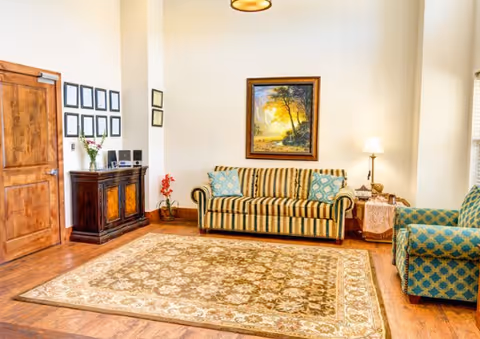 A cozy living room with a striped sofa and a patterned armchair, a large decorative rug on wooden flooring, a wooden cabinet with framed pictures and flowers, a floor lamp beside the sofa, and a framed painting of a forest scene on the wall.