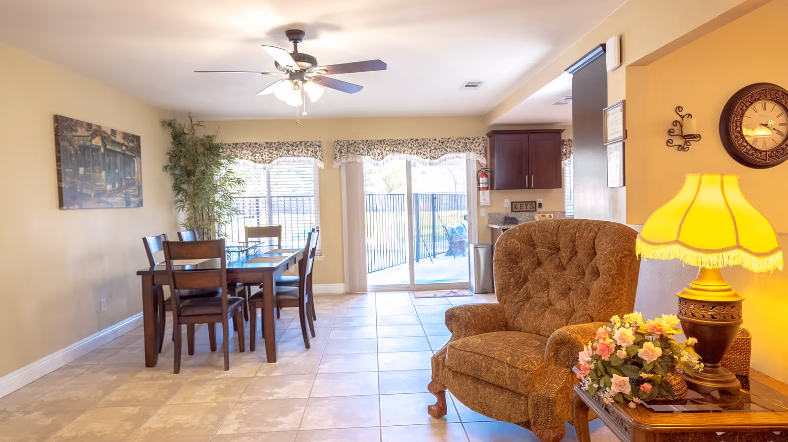 A bright and cozy living and dining area in Annie's Haven featuring a wooden dining table with six chairs, a large window with floral valances, a sliding glass door leading outside, a comfortable upholstered armchair, a side table with a yellow lamp and a flower arrangement, a ceiling fan with lights, and a wall clock.