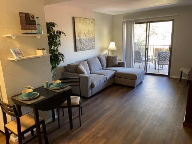 A cozy living room with a gray sectional sofa, a floor lamp, and a large window with sliding glass doors leading to a balcony with outdoor chairs. There is a small dining table with two place settings and chairs near the left side, along with wall shelves holding decorative items and a potted plant in the corner.