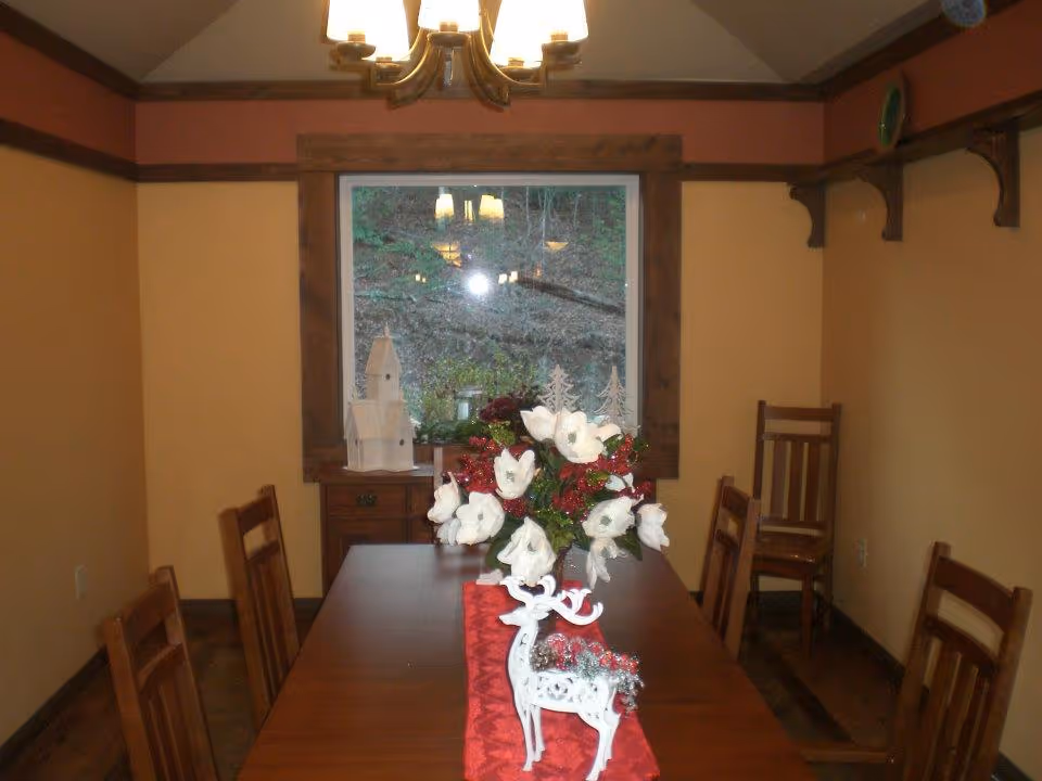 A dining room with a wooden table and six wooden chairs. The table is decorated with a red runner, a white decorative reindeer, and a floral centerpiece with white flowers and red berries. There is a window with a wooden frame at the end of the room, showing an outdoor view. A chandelier with multiple lights hangs from the ceiling above the table. The walls are painted a warm beige color with wooden trim and a shelf on the right side.