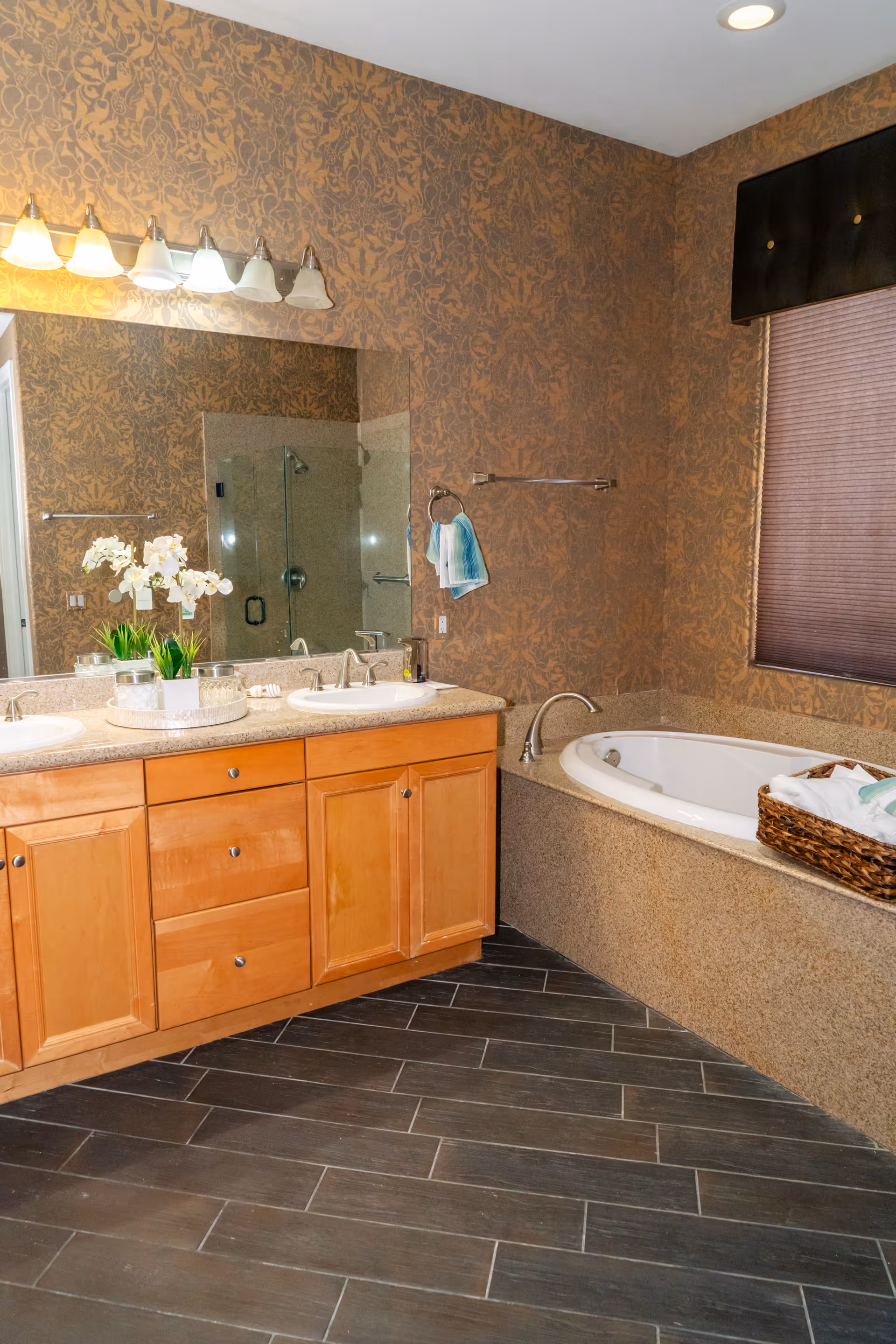 A bathroom with a double sink vanity featuring wooden cabinets and a large mirror above. There is a bathtub with a granite surround to the right, a basket with towels on the tub edge, and a glass-enclosed shower reflected in the mirror. The walls have a brown patterned wallpaper, and the floor is covered with dark wood-like tiles. A window with a dark shade is above the bathtub.