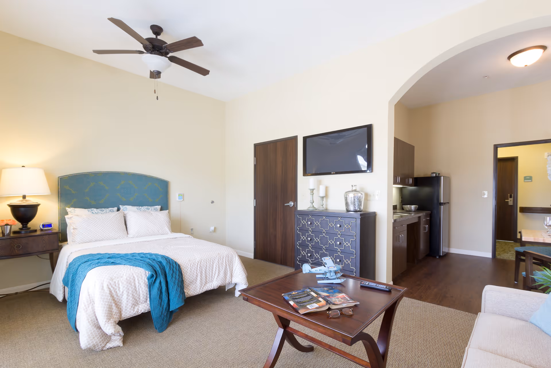 A bright and cozy senior living bedroom with a bed featuring a blue upholstered headboard and white bedding with a teal throw blanket. There is a wooden nightstand with a lamp and clock, a ceiling fan above, a wooden coffee table with books and a model airplane, a decorative cabinet with candles and a vase, a wall-mounted TV, and a kitchenette visible through an archway.