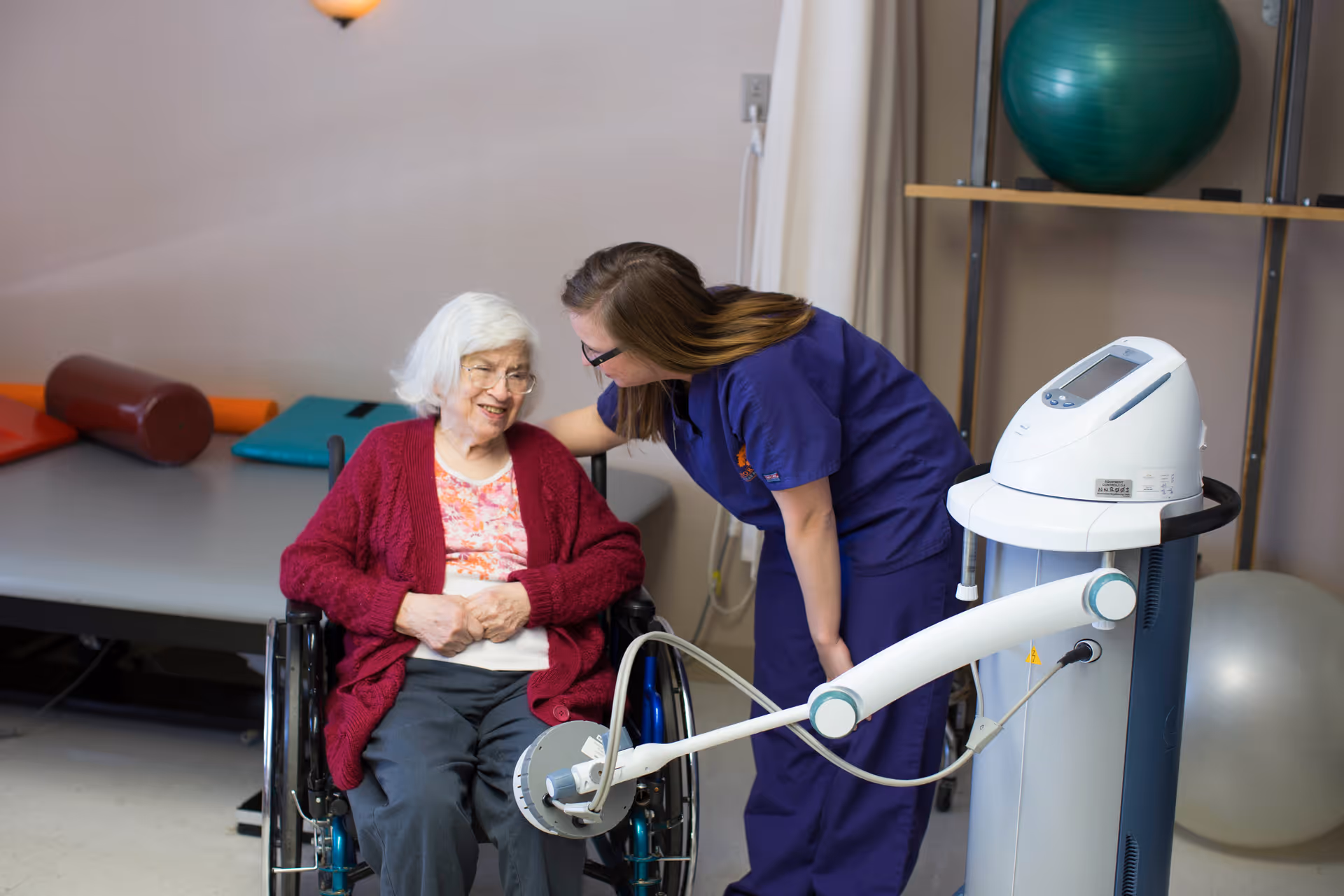An elderly woman in a wheelchair wearing a red cardigan and floral shirt smiles while a healthcare worker in purple scrubs leans toward her, smiling and engaging. They are in a room with physical therapy equipment including a large exercise ball and a medical device with an arm extending toward the woman.