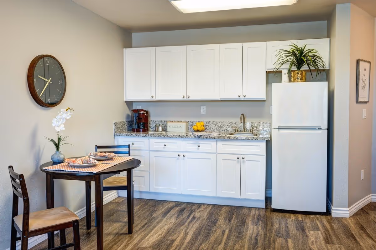 A small kitchen area with white cabinets, a granite countertop, a white refrigerator, and a stainless steel sink. A wooden dining table with two chairs is set with placemats, plates, and a vase with white flowers. A round wall clock is mounted on the beige wall above the table. The floor has wood-style vinyl flooring.