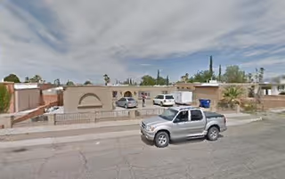 Street view of a single-story residential building with a beige exterior and a low wall in front. A silver pickup truck is parked on the street in front of the building. The sky is partly cloudy.