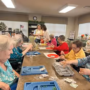 A group of elderly women seated around a large table engaged in a craft or activity session inside a well-lit room with windows and shelves in the background. Two women are standing and assisting others. Various craft materials and trays are on the table.