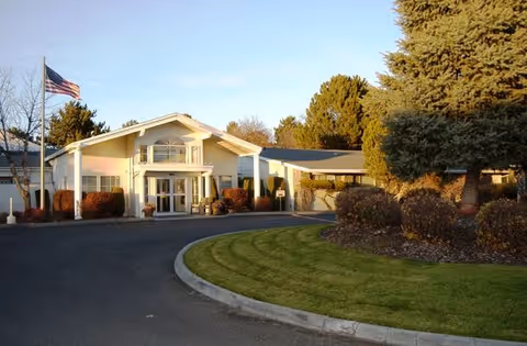 Exterior view of a single-story building with a peaked roof and large windows, surrounded by neatly trimmed bushes and trees. An American flag is flying on a flagpole to the left of the building. A curved driveway leads up to the entrance, and the area is well-maintained with green grass and landscaping.