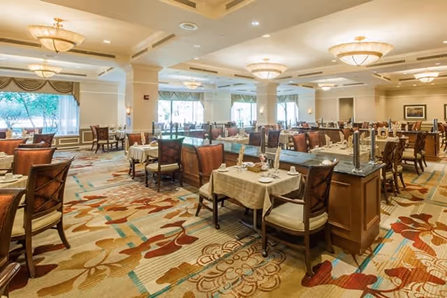 A spacious and well-lit dining room with multiple tables covered in white tablecloths, set with plates and napkins. The room features large windows with curtains, patterned carpet, and elegant ceiling lights.