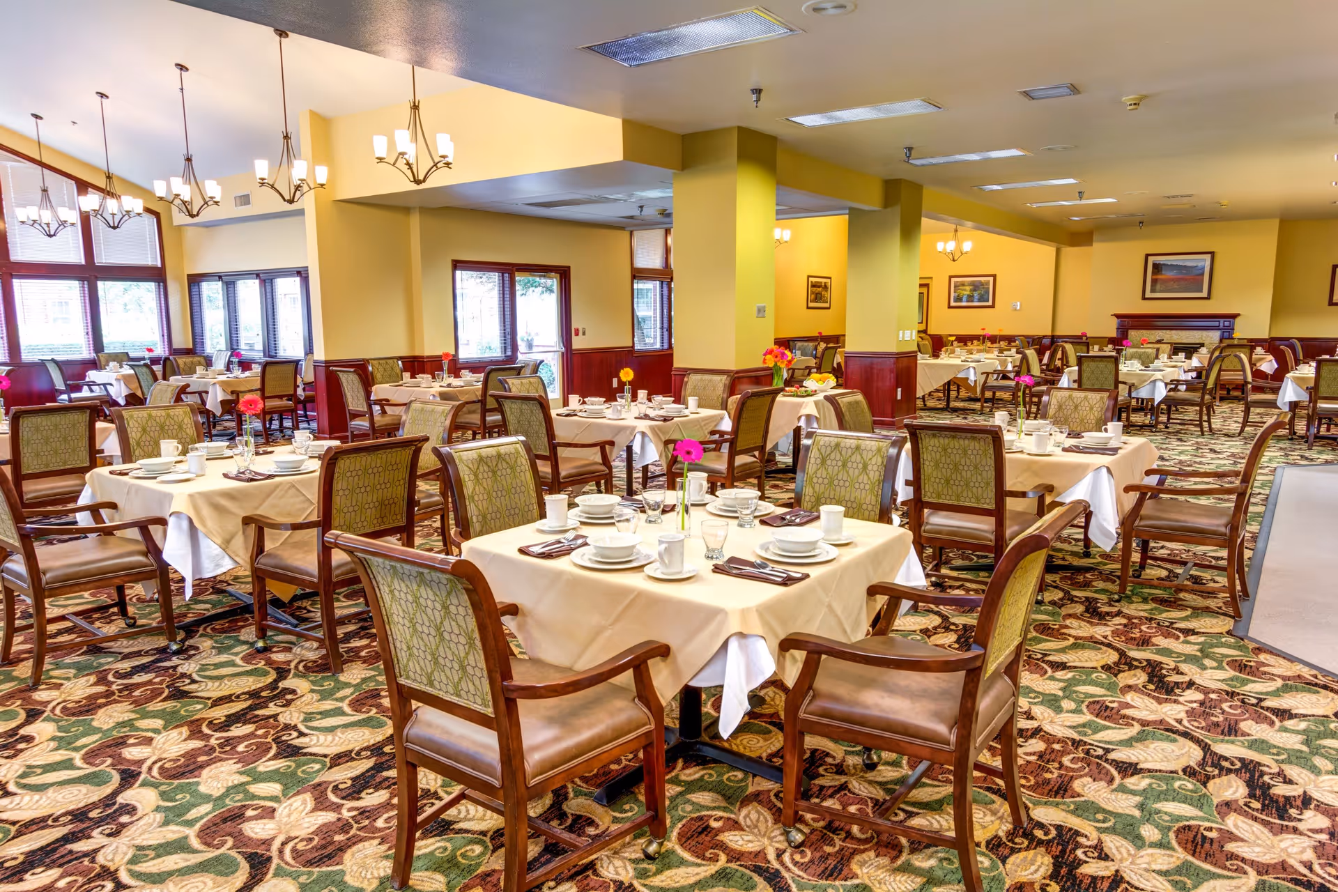 A spacious dining room with multiple tables covered in beige tablecloths, each set with white dishes, cups, and silverware. The room features patterned carpet, wooden chairs with green upholstery, yellow walls, and several chandeliers hanging from the ceiling. Large windows allow natural light to fill the space.