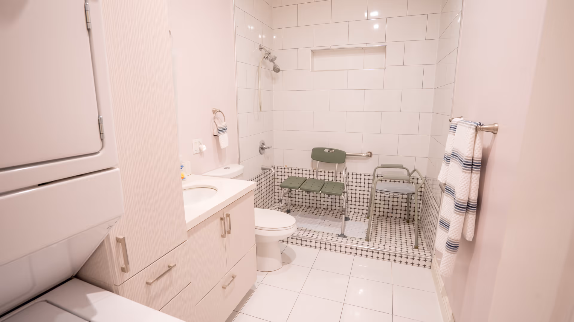 A bathroom with a white tiled walk-in shower equipped with a green shower chair and a green stool. The bathroom also features a white toilet, a vanity with a sink, a towel ring with a white and blue striped hand towel, and a stacked washer and dryer unit. Another towel with blue stripes hangs on a towel bar on the right wall.