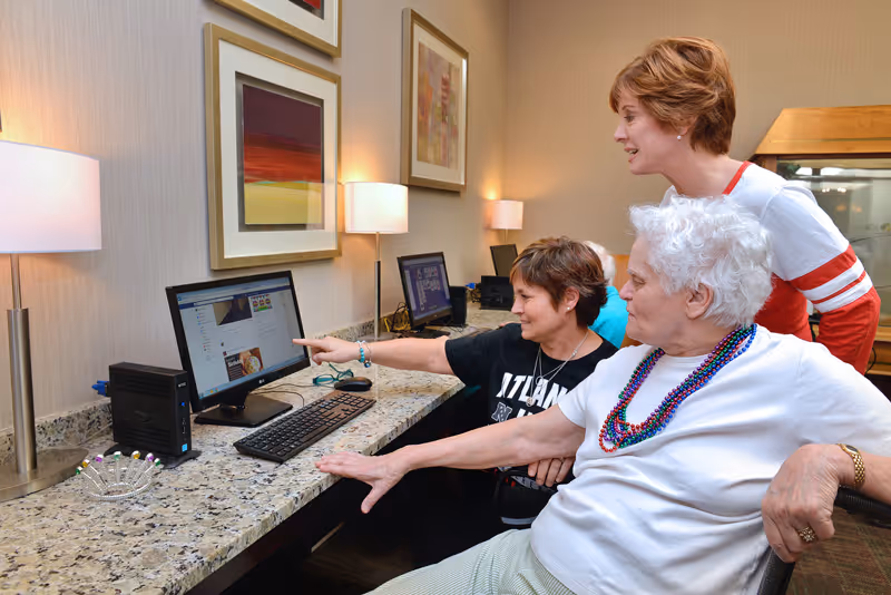 Three women gathered around a computer in a well-lit room with granite countertops and framed artwork on the wall. One woman is seated in a wheelchair wearing colorful bead necklaces, another woman is pointing at the computer screen, and a third woman is standing behind them, smiling and observing.