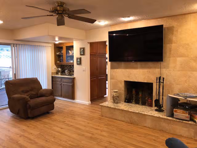 Spacious living room with hardwood floors, a wall-mounted TV above a tiled fireplace, a brown recliner, ceiling fan, and sliding glass doors.
