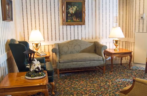 A cozy living room area with a green upholstered loveseat centered against a wallpapered wall. On either side of the loveseat are wooden side tables with brass lamps emitting warm light. To the left, there is a dark green armchair and a wooden table with a decorative floral arrangement. A framed painting of flowers hangs above the loveseat. The floor is covered with a patterned carpet.