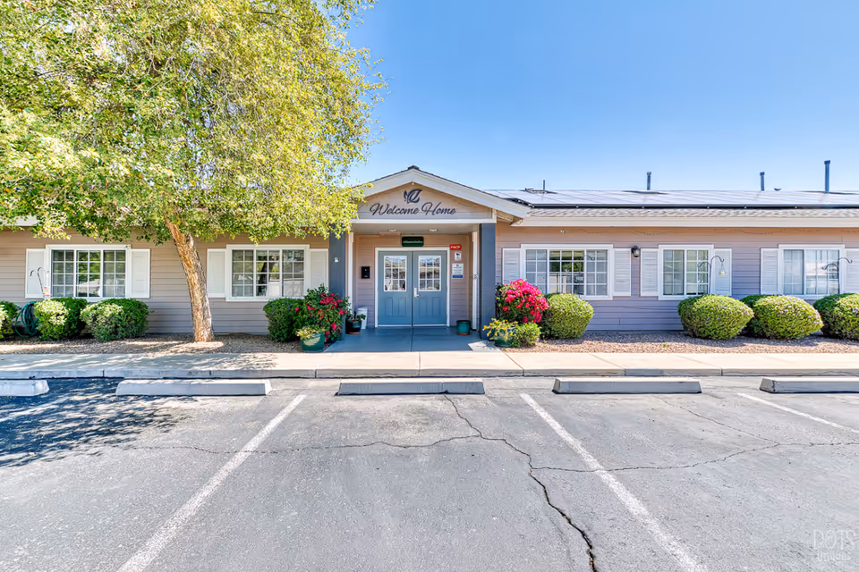 Exterior front view of Paradise Valley Senior Living facility showing a single-story building with a welcoming entrance. The building has light gray siding, white window shutters, and a sign above the door that reads 'Welcome Home'. There are green bushes and colorful flowers planted along the front, and a large tree providing shade on the left side. The parking area in front has several empty parking spaces under a clear blue sky.