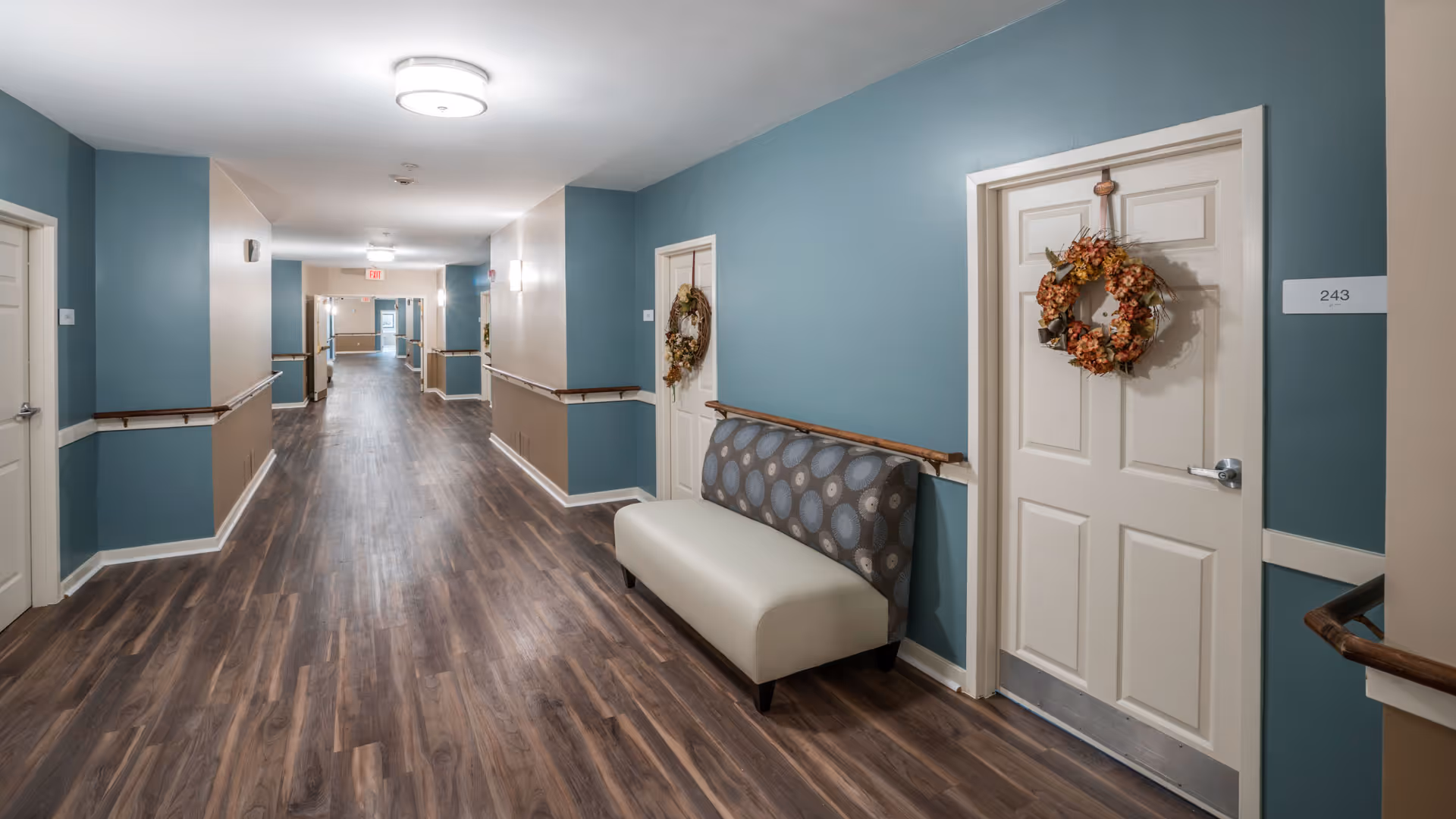 A long, well-lit hallway in a senior living facility with wood-patterned flooring and teal and beige walls. Several doors line the hallway, each decorated with a fall-themed wreath. A cushioned bench with a patterned backrest is placed against the wall near one of the doors labeled 243. Handrails run along both sides of the hallway.
