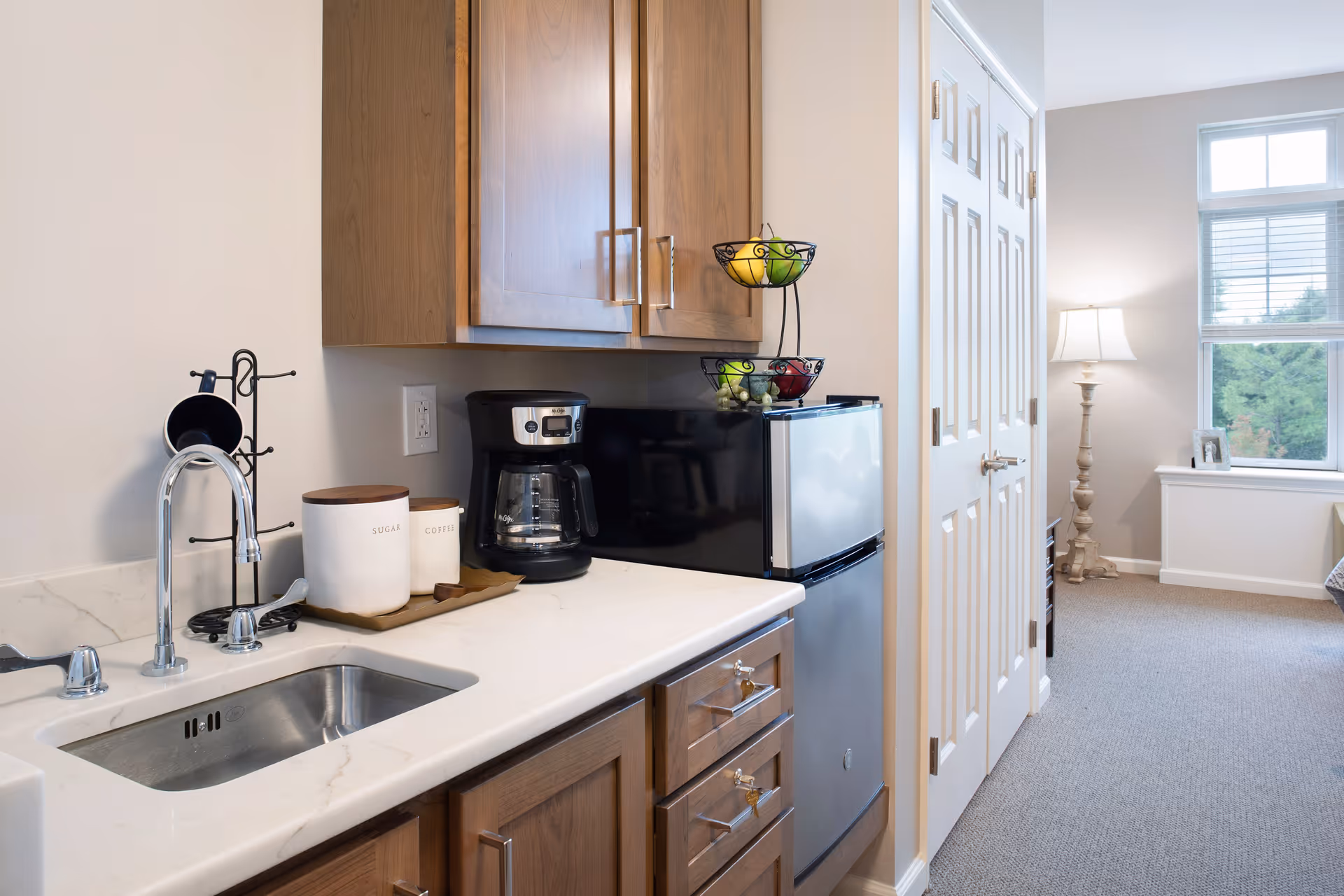 A small kitchenette area with a stainless steel sink, a coffee maker, two containers labeled sugar and coffee on a tray, and a mini refrigerator topped with a fruit basket. The kitchenette has wooden cabinets and drawers. In the background, there is a carpeted room with a window, a lamp on a stand, and a small framed photo on the windowsill.