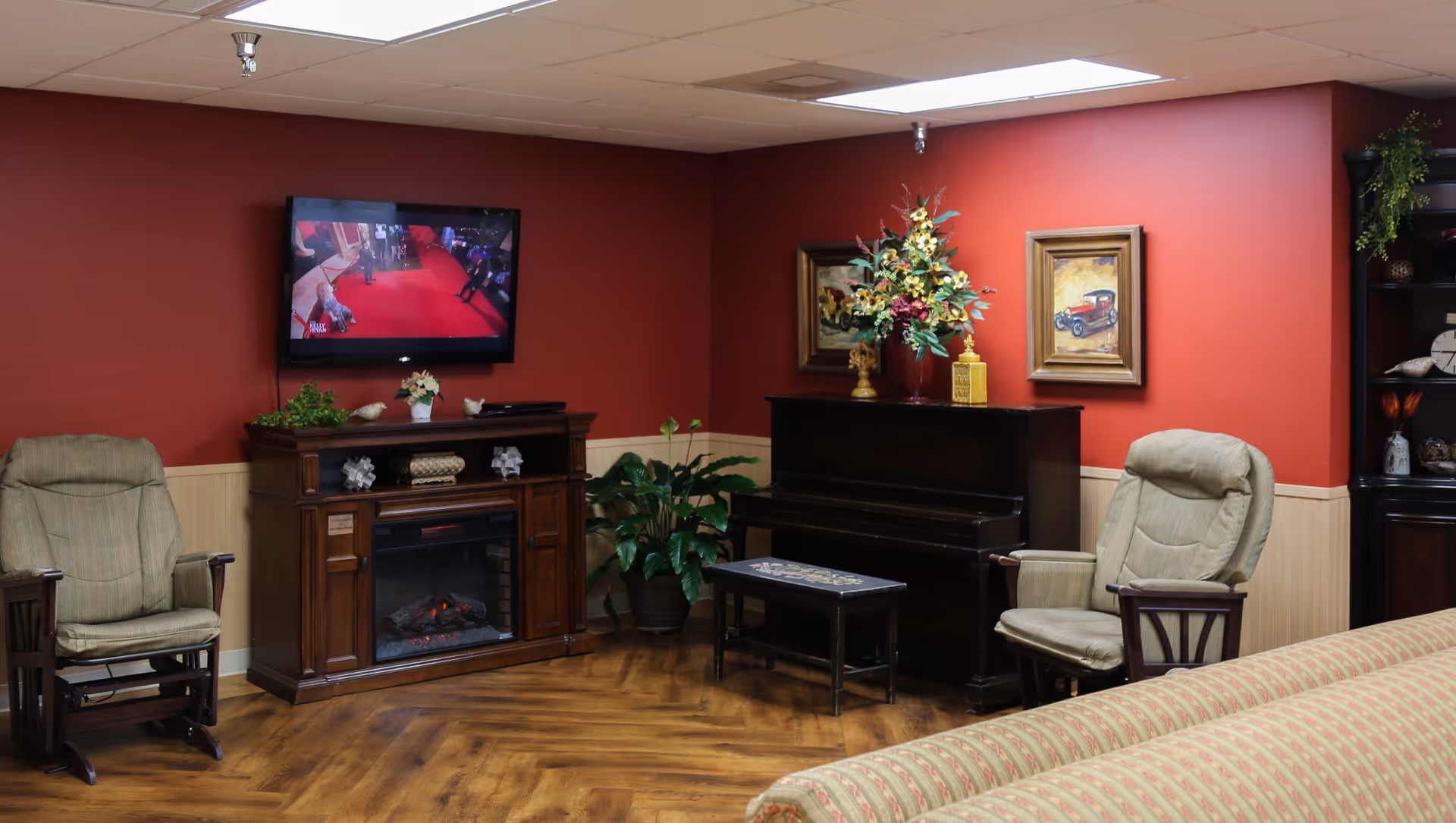 A cozy living room with red walls and wood paneling. The room features two cushioned armchairs, a wooden electric fireplace with decorative items on top, a flat-screen TV mounted on the wall showing a red carpet event, a dark wooden piano with a floral arrangement on top, framed paintings on the wall, a potted plant, and a striped sofa partially visible in the foreground.