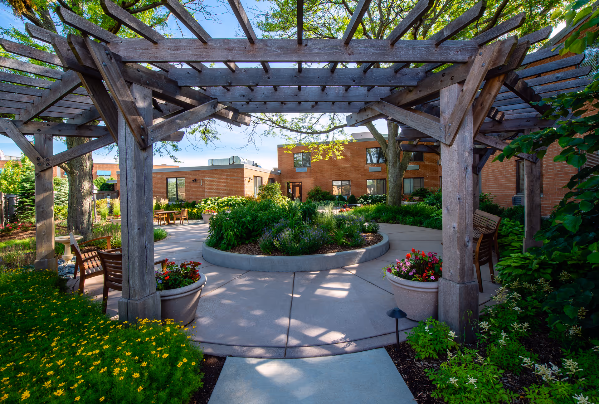 Outdoor garden area at Central Baptist Village featuring a wooden pergola, circular concrete pathway, lush green plants, colorful flowers in large pots, wooden benches, and brick buildings in the background under a clear blue sky.