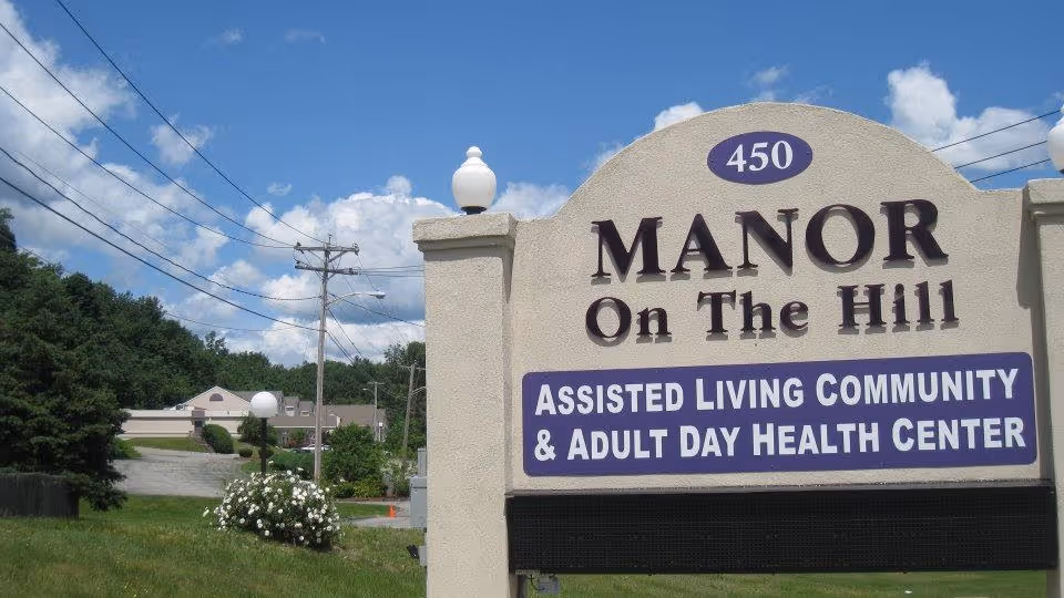 Large entrance sign reading "Manor On The Hill" for an assisted living community with the facility and trees behind it under a blue sky.