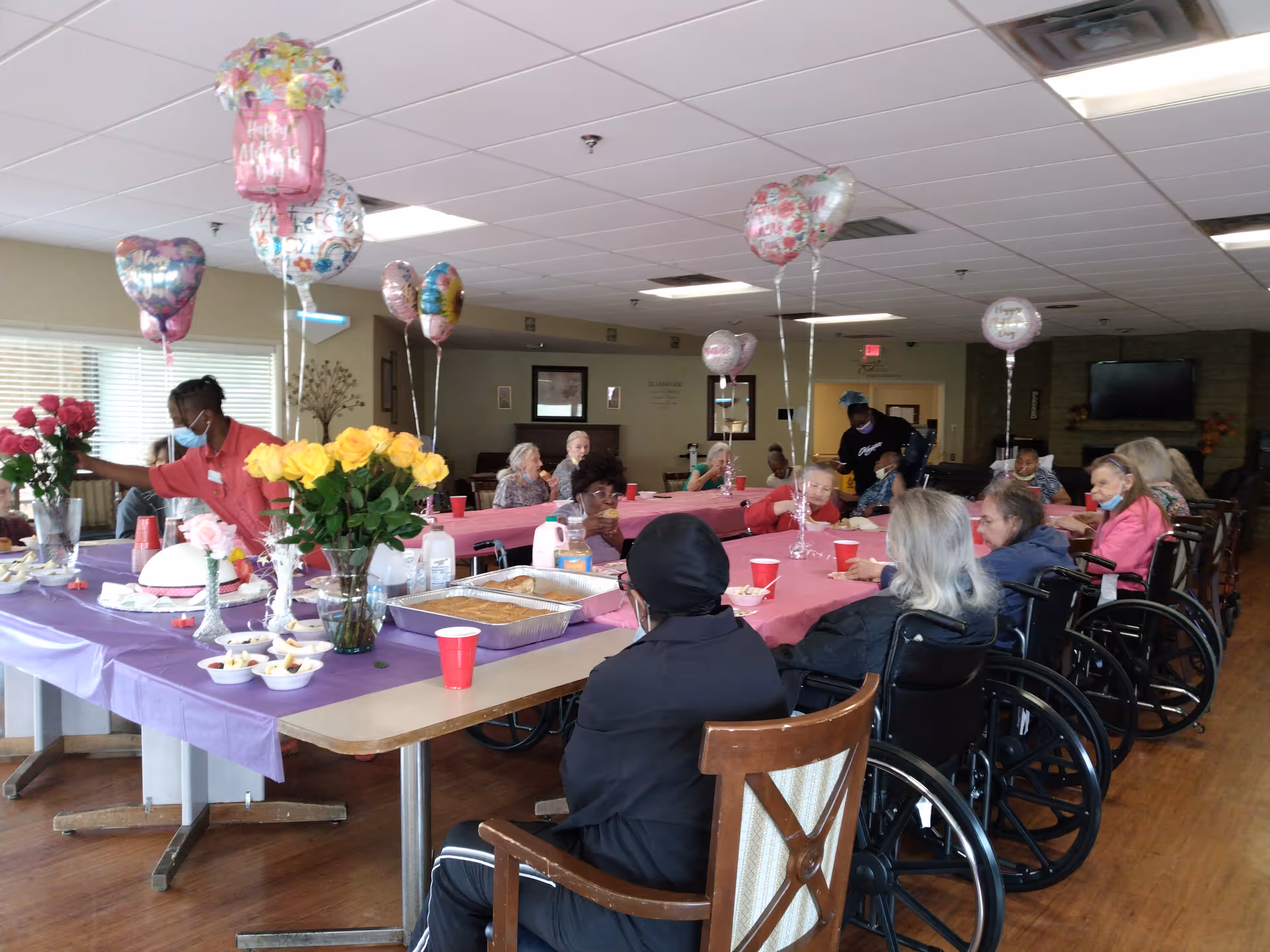 A group of elderly people, many in wheelchairs, seated around tables covered with pink and purple tablecloths in a communal dining area. The tables are decorated with balloons and vases of flowers. A staff member wearing a mask is arranging flowers on one table. The room has a warm, welcoming atmosphere with wooden floors and a television mounted on the wall in the background.