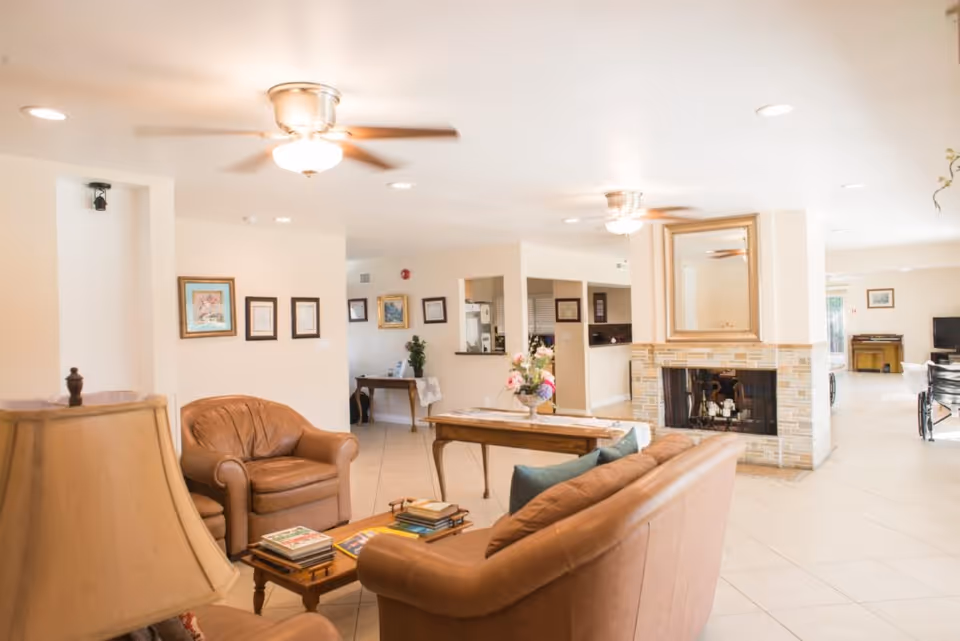 Bright communal living room with leather sofas and armchairs arranged around a central table and fireplace with ceiling fans overhead.