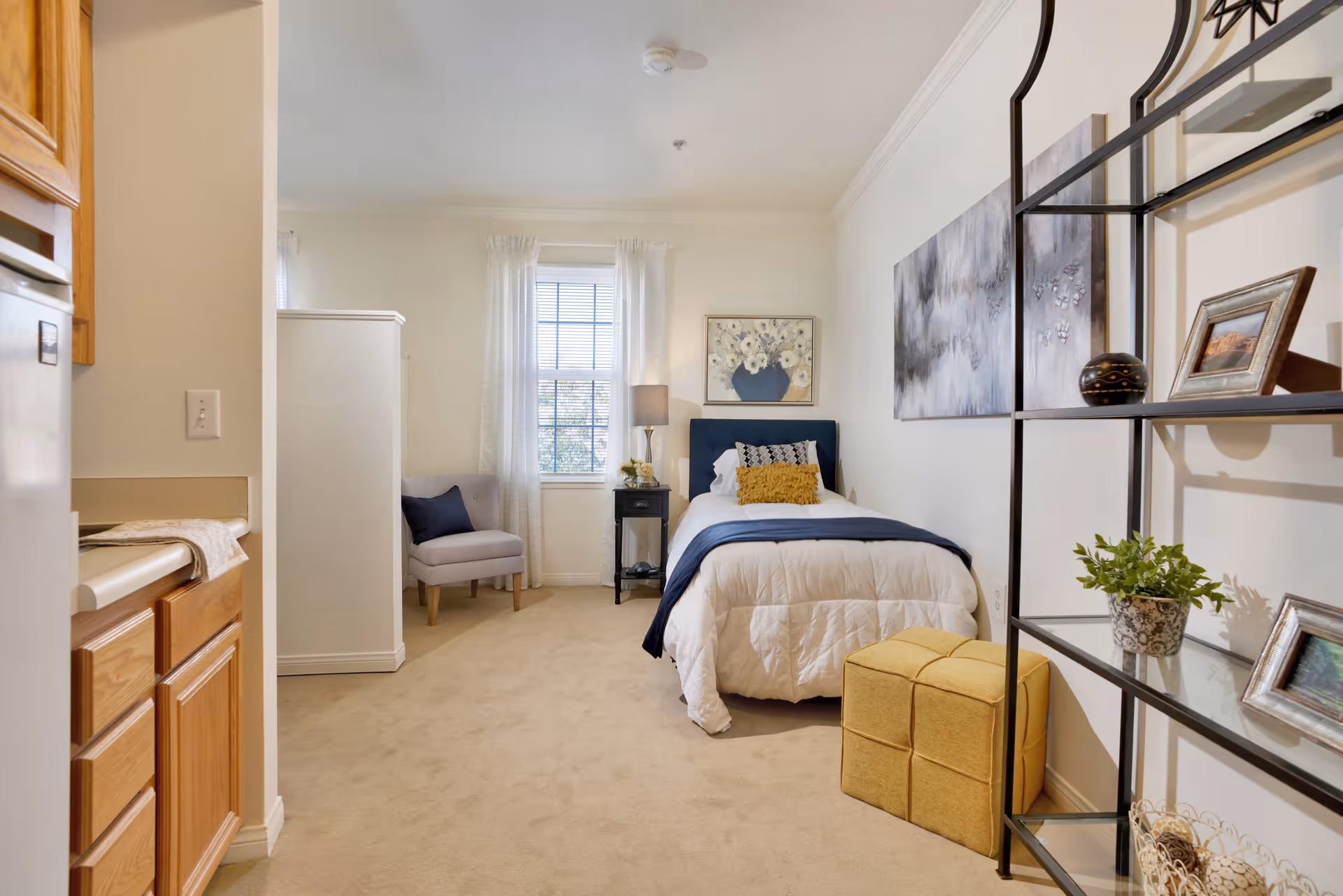 A cozy bedroom with a single bed dressed in white and navy bedding, a yellow cushion, and a nightstand with a lamp and flowers. There is a window with sheer white curtains, a gray armchair with a navy pillow, a yellow ottoman, and a black metal shelving unit with framed photos and a plant. The room has beige carpet and light-colored walls with artwork hanging above the bed.