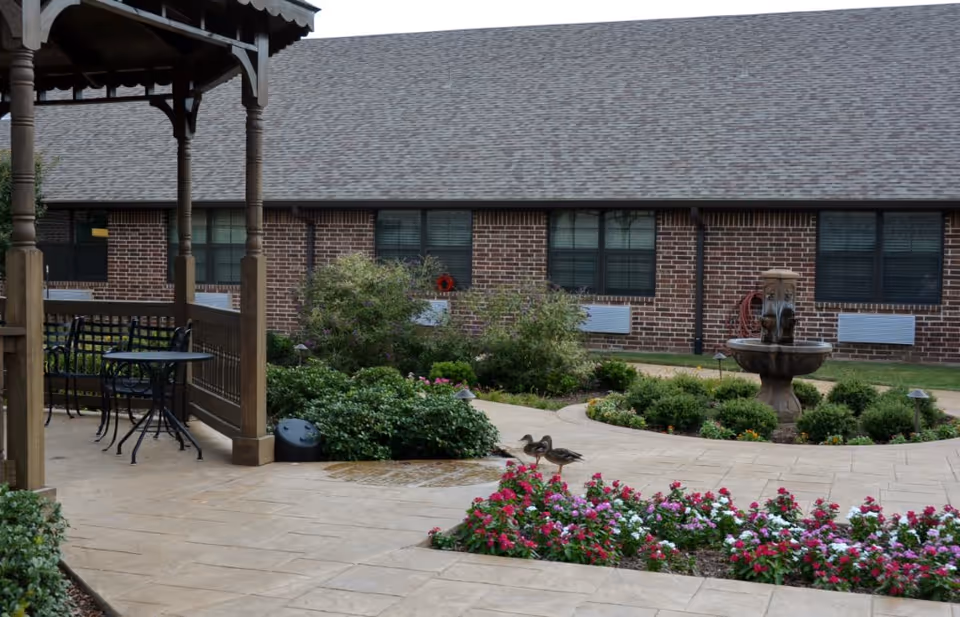 Outdoor courtyard area with a gazebo featuring a table and chairs, surrounded by flower beds with colorful flowers, bushes, a stone fountain, and a brick building in the background. Two ducks are walking on the paved pathway near the flower beds.