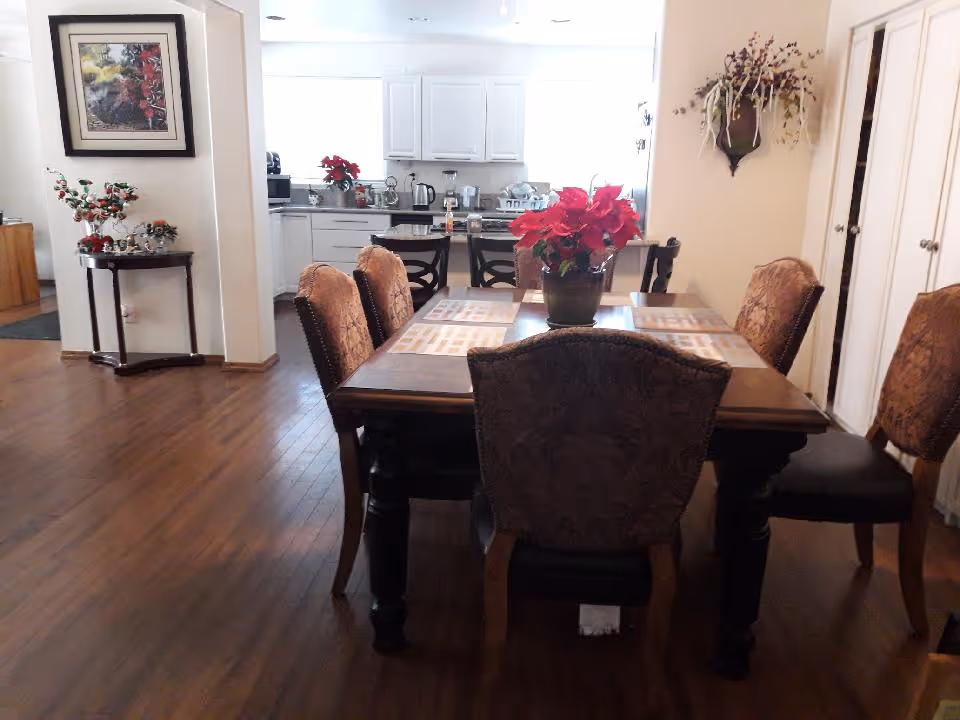 Dining area with a wooden table set for six with placemats and a red poinsettia centerpiece. The room has hardwood floors and is adjacent to a kitchen with white cabinets and appliances. Decorative items include a framed picture on the wall, a small table with holiday decorations, and a wall-mounted floral arrangement.