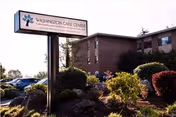 Exterior view of Washington Care Center building with a large sign displaying the facility's name. The scene includes landscaped bushes and trees with a clear sky in the background.