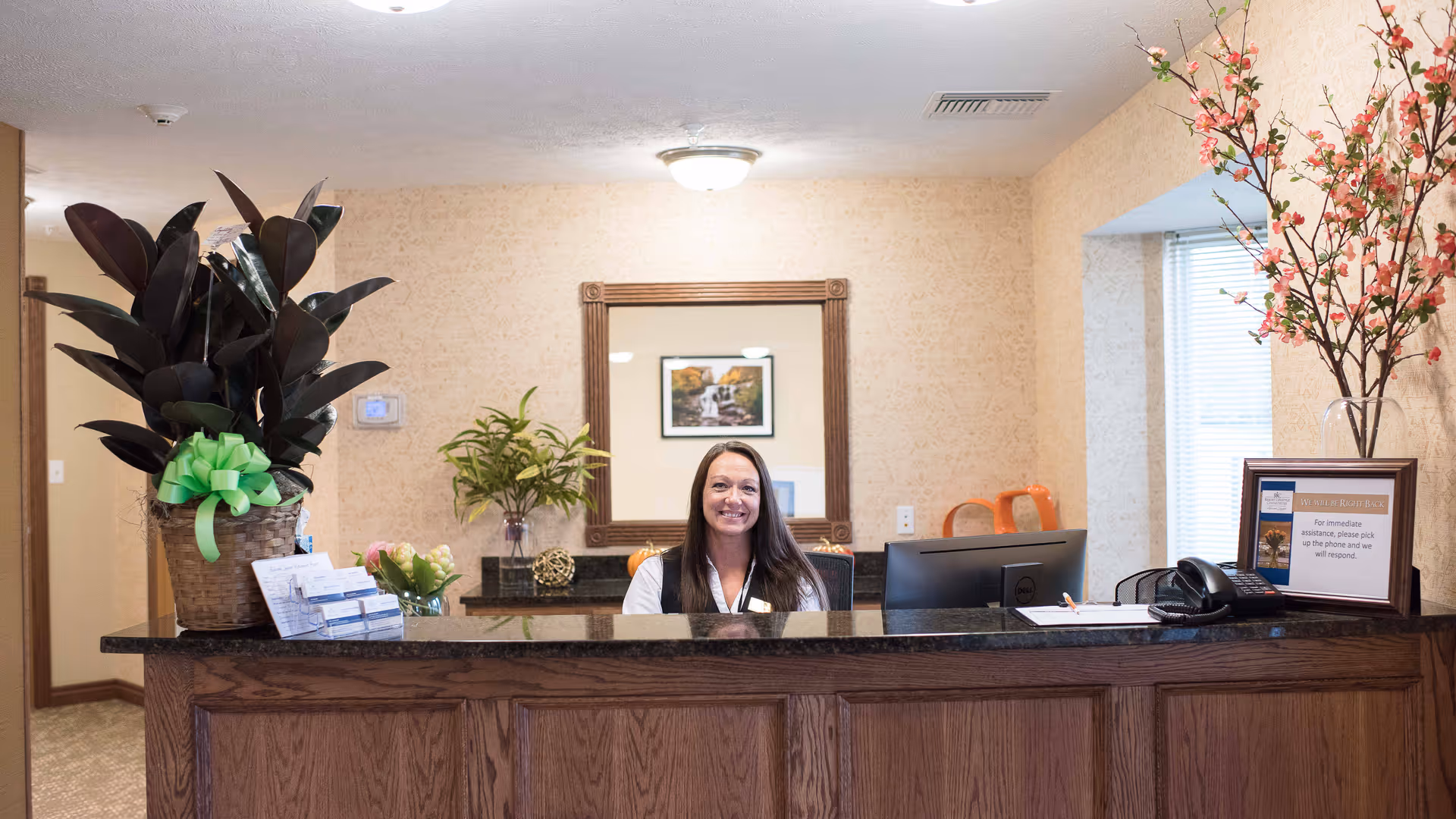 Reception desk area at Ashwood Square Retirement Community with a smiling receptionist seated behind a dark granite countertop. The desk has a large potted plant with a green bow, a small bouquet of flowers, brochures, a computer monitor, a phone, and a framed sign. Behind the receptionist is a wall mirror reflecting a framed picture and some decorative plants. The walls are beige with a subtle pattern, and there is a window with blinds on the right side.