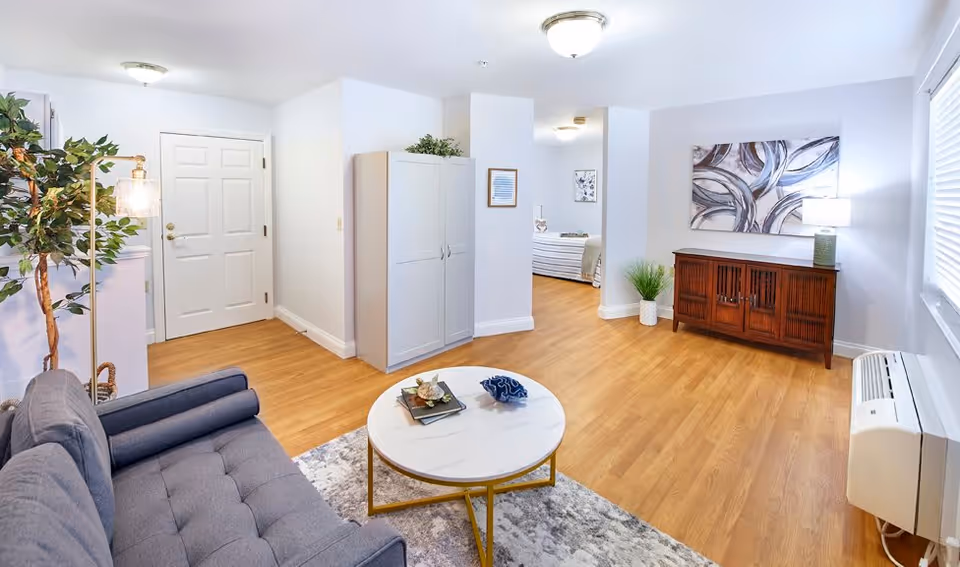 A bright and spacious living room with light wood flooring, a gray tufted sofa, a round white marble coffee table with gold legs, a wooden cabinet with a lamp and abstract artwork above it, a potted plant, and a large window with blinds. A doorway leads to a bedroom with a visible bed and framed pictures on the walls.