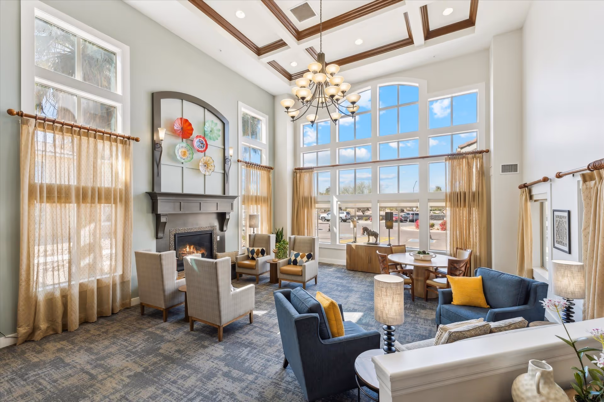 A bright and spacious living room area with large windows letting in natural light. The room features a fireplace with decorative plates above it, several armchairs and sofas with cushions, a round table with chairs near the windows, and elegant curtains. A chandelier hangs from the ceiling, and there are lamps and plants adding to the cozy atmosphere.