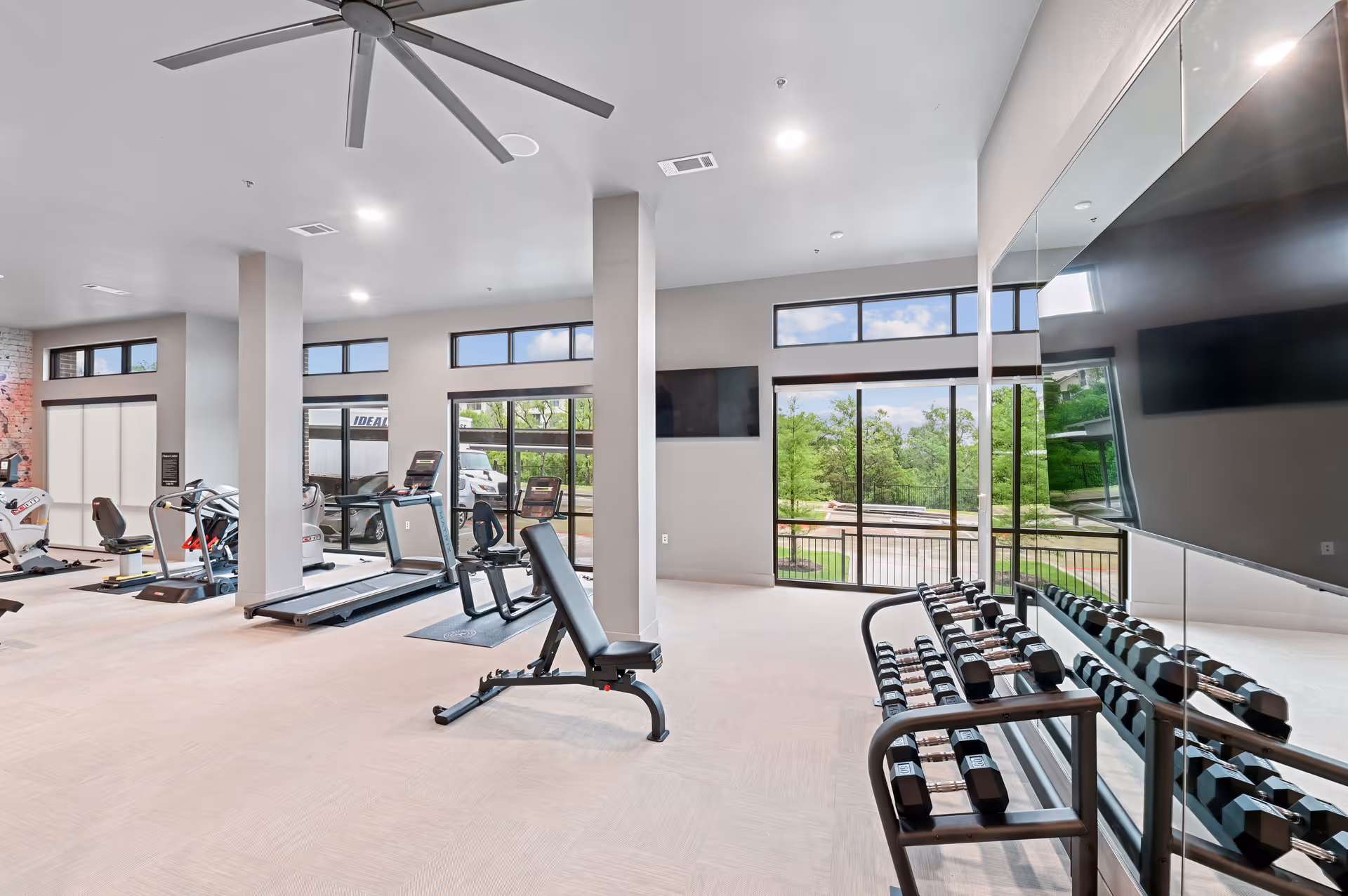 Bright and spacious fitness room with exercise equipment including treadmills, stationary bikes, a weight bench, and a rack of dumbbells in front of a large mirror. Large windows provide natural light and a view of greenery outside.