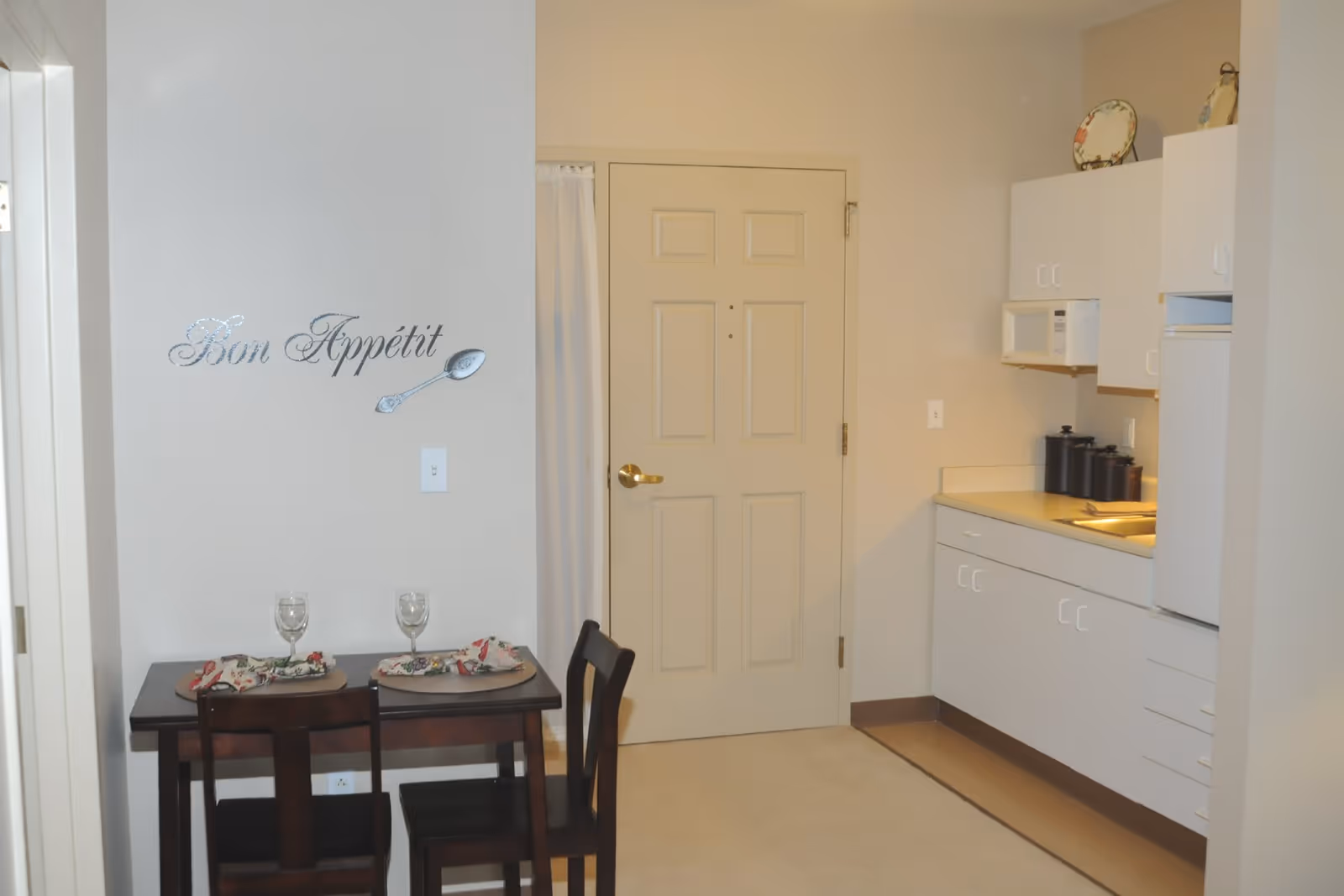 Small apartment entry with a kitchenette on the right and a two-seat dining table beneath a 'Bon Appétit' wall decal.