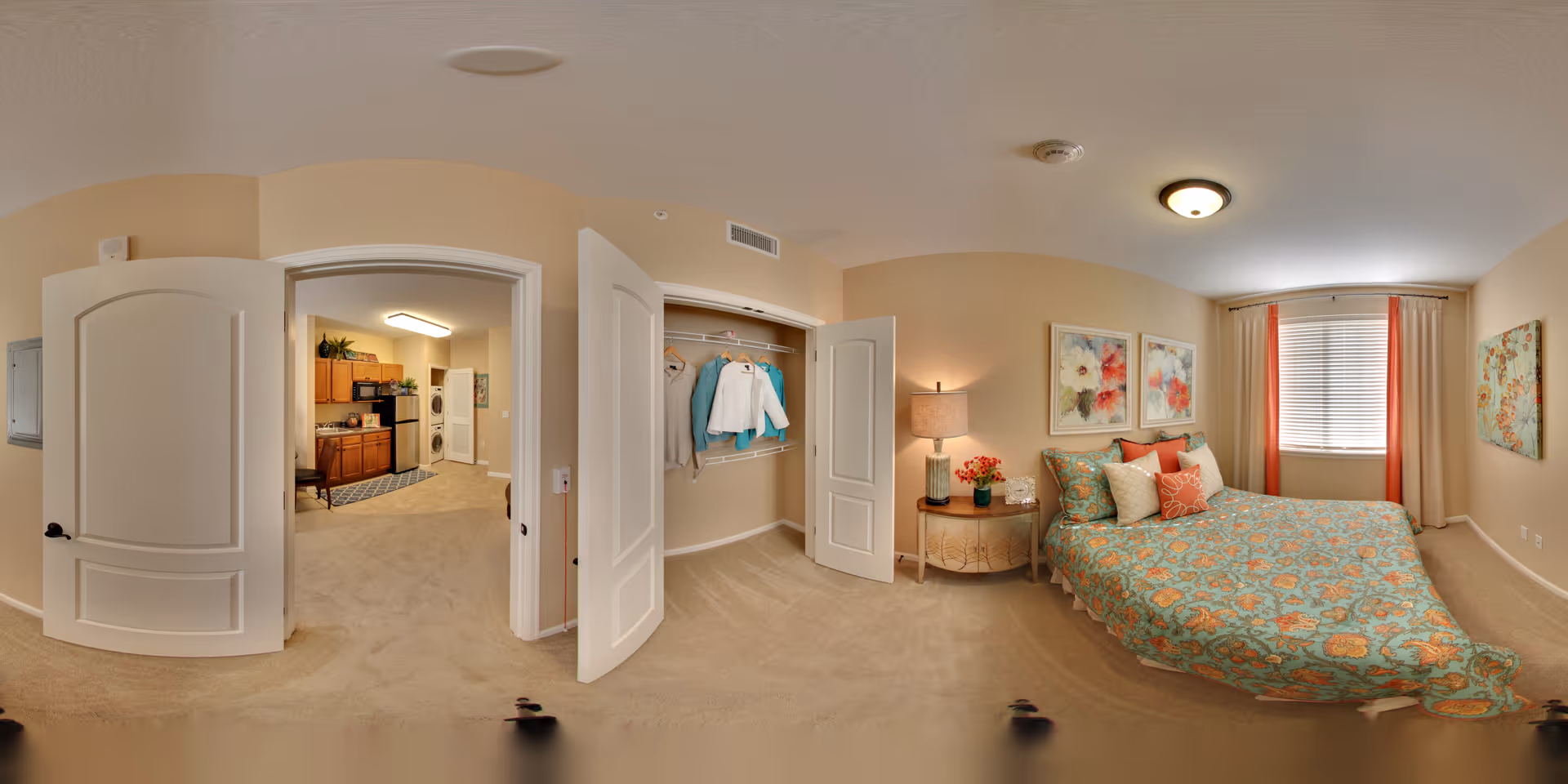 A cozy bedroom in an assisted living facility with a bed covered in a colorful floral bedspread and multiple pillows. There is a nightstand with a lamp and flowers next to the bed. The room has beige walls, a window with blinds and coral-colored curtains, and floral artwork on the walls. An open closet with clothes hanging inside is visible, as well as an open door leading to a kitchen area with wooden cabinets and appliances.