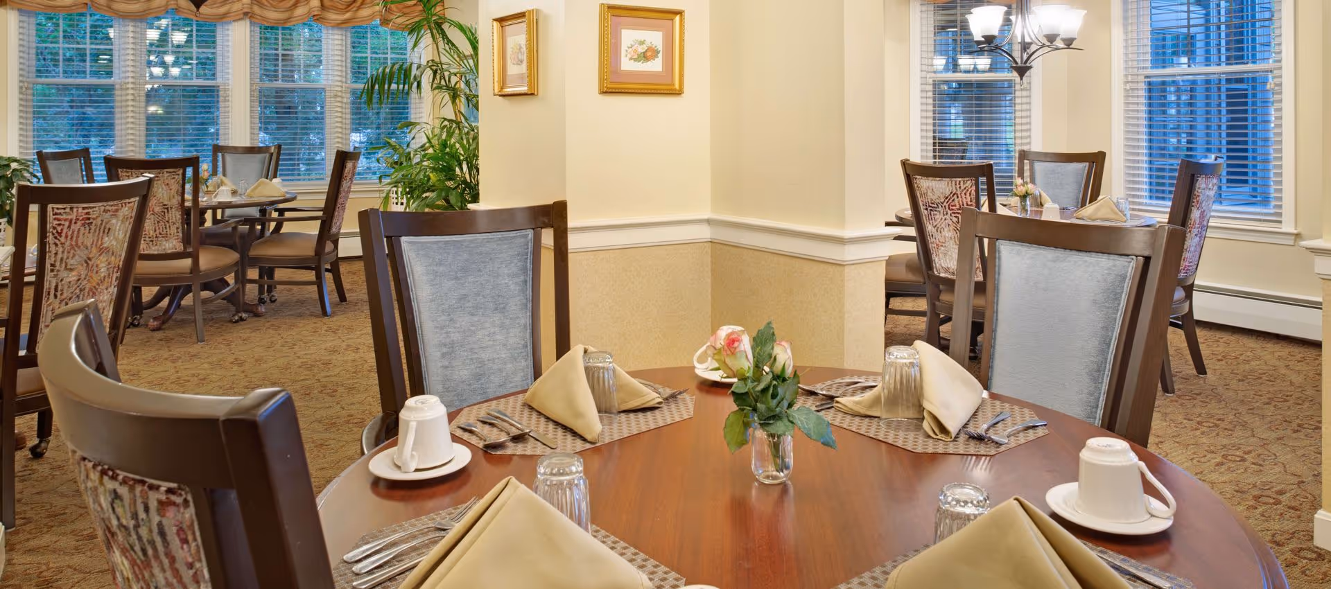 A dining room with round wooden tables set with beige napkins, glasses, cups, and silverware. The room has large windows with blinds, patterned carpet, and framed floral artwork on the walls. There are upholstered chairs around the tables and a chandelier hanging from the ceiling.
