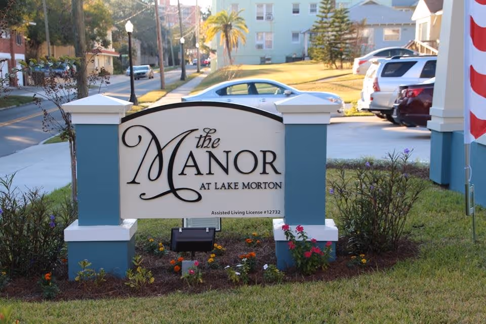 Outdoor view of a sign for The Manor at Lake Morton assisted living facility, surrounded by small plants and flowers, with a street and parked cars in the background.