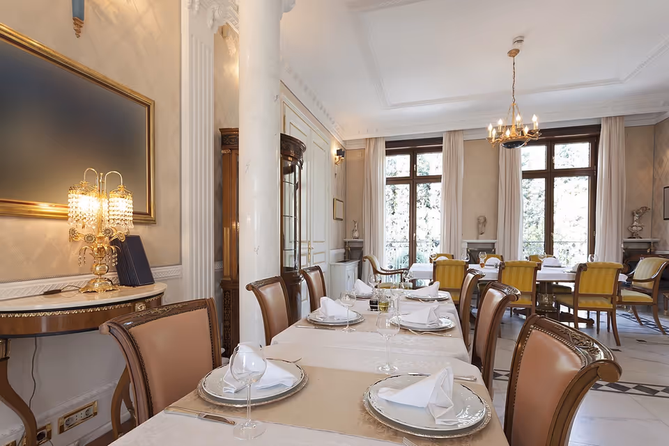 Elegant dining room with a long table set for a meal, featuring white tablecloths, neatly folded napkins, wine glasses, and silver plates. The room has large windows with white curtains, classic wooden chairs with leather upholstery, ornate wall decorations, a chandelier, and a decorative lamp on a side table.